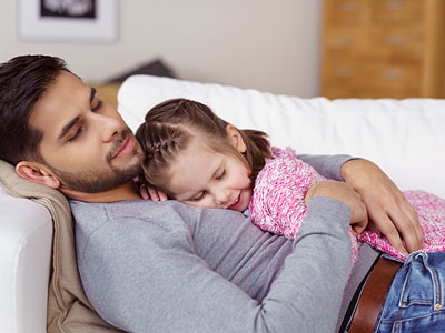 Ein Vater und sein Kind liegen gemütlich auf einer Couch. Das Kind liegt schlafend im Arm des Vaters.